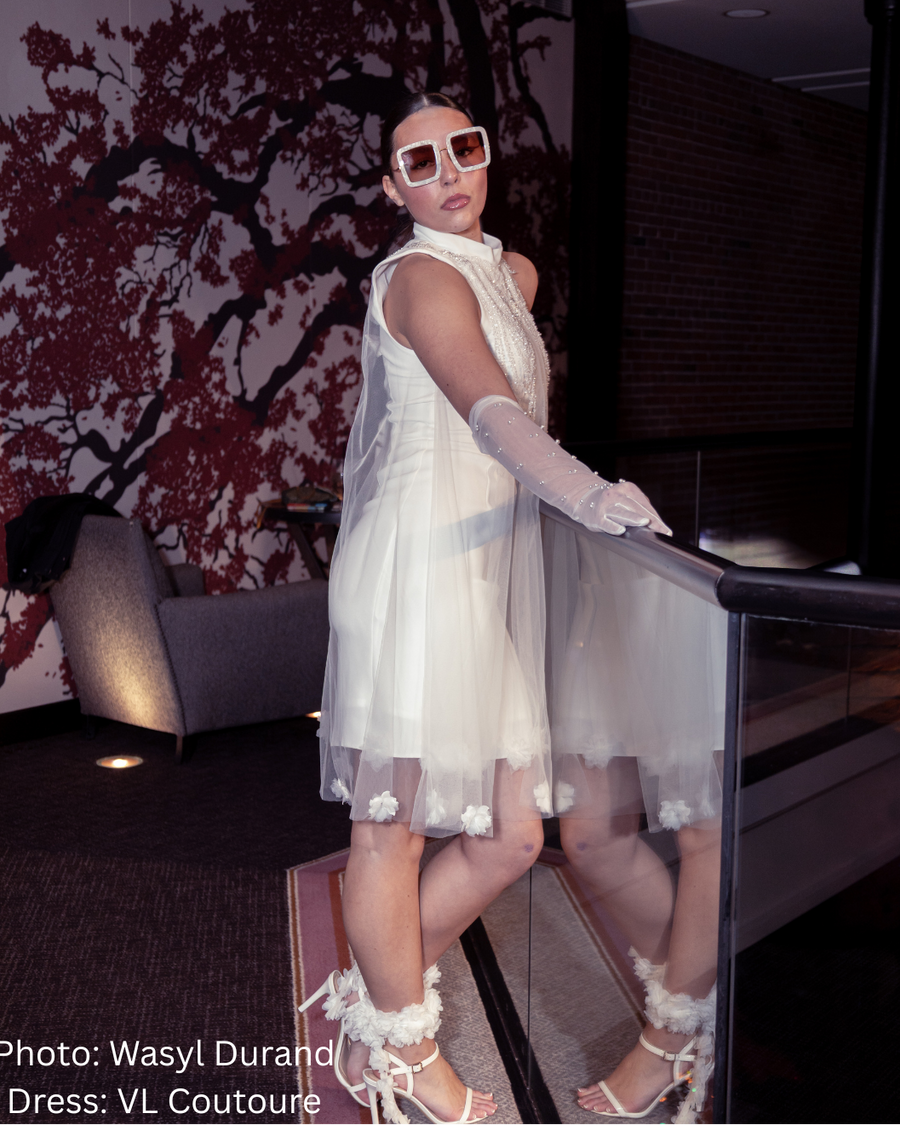 Woman in a wedding dress and sunglasses standing indoors with a decorative wall in the background.